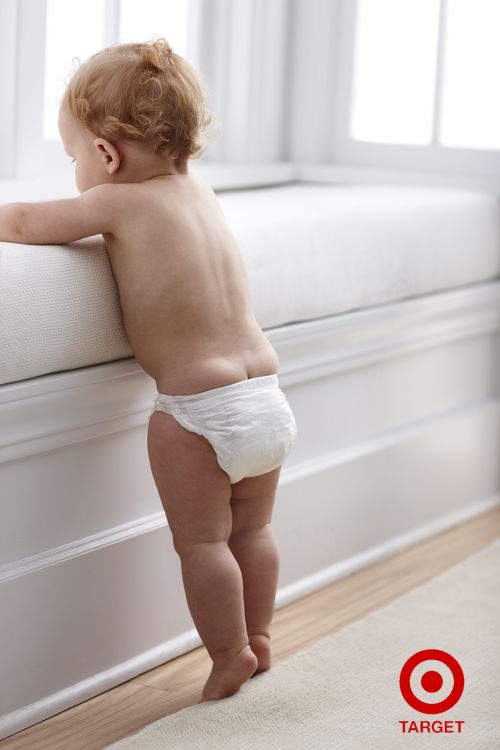 Infant wearing a white diaper is standing and holding onto a white cushioned bench in front of a white wall and light carpet.