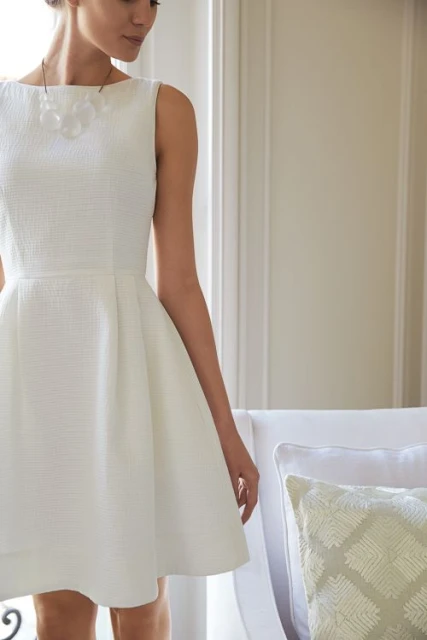 Adult female poses in a white textured A-line dress and necklace. The image features bright indoor lighting and a neutral color palette.