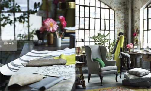 Interior design featuring grey wingback chair, textured fabrics, pillows, and tables, set in a sunlit room with large windows.