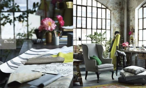 Interior design featuring grey wingback chair, textured fabrics, pillows, and tables, set in a sunlit room with large windows.