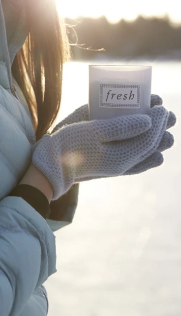 Woman in gloves holds 'fresh' candle outdoors in a bright, snowy environment with sunlight, creating a serene winter atmosphere.