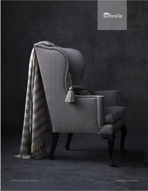Wingback chair with a grey striped throw blanket, decorative tassel, and cushion in a dimly lit room with a textured wall.