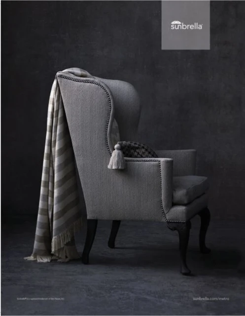 Wingback chair with a grey striped throw blanket, decorative tassel, and cushion in a dimly lit room with a textured wall.
