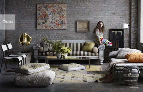 A young woman sits on a grey couch with striped cushions in a room with a grey brick wall, patterned rug and cushions, and chairs.
