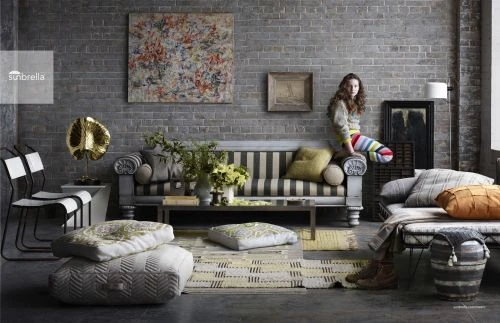 A young woman sits on a grey couch with striped cushions in a room with a grey brick wall, patterned rug and cushions, and chairs.