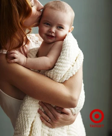 A baby wrapped in a white blanket being held by a woman with red hair, with the Target logo in the bottom right corner.