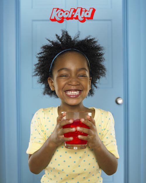 Smiling African-American girl holds glass of red drink in front of a light blue door with Kool-Aid logo above her head.