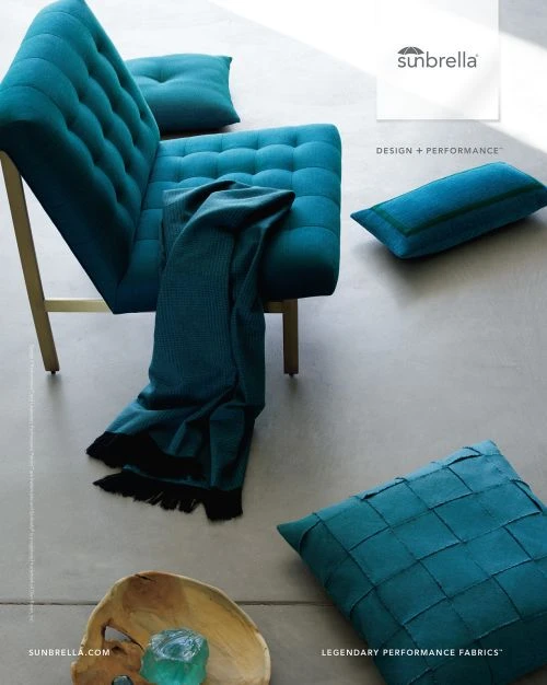 Studio shot of a modern teal fabric sofa and matching pillows arranged on a grey floor with a wooden bowl and glass cube.