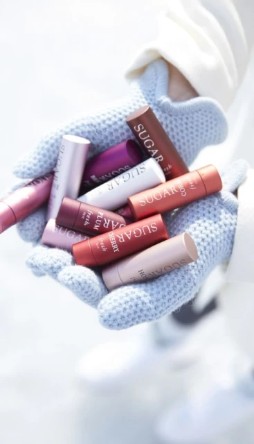 Closeup of a person holding a collection of colorful Sugar lip balms in their gloved hands against a blurred light background.