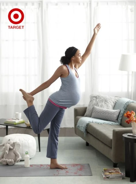 Pregnant woman in gray tank and yoga pants performs yoga pose on yoga mat inside a bright room with couch and pillows.