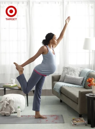 Pregnant woman in gray tank and yoga pants performs yoga pose on yoga mat inside a bright room with couch and pillows.