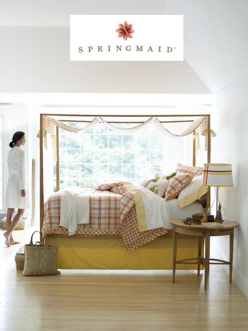 Bright and airy bedroom interior featuring a four-poster bed with plaid bedding, side table, and a woman in a robe walking by.
