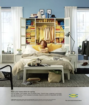 Bedroom interior showcasing a made bed, open wardrobe with colorful clothing, and a woman resting with yellow pillows against a blue wall.