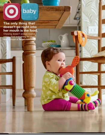 Baby sitting on a wooden floor biting a colorful plush toy dog in a domestic kitchen setting