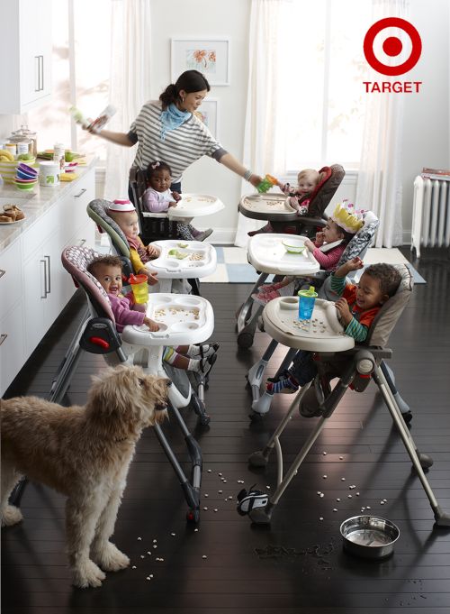Six infants and one adult female caregiver, surrounded by white cabinetry in a bright, indoor kitchen, with a tan-colored dog.