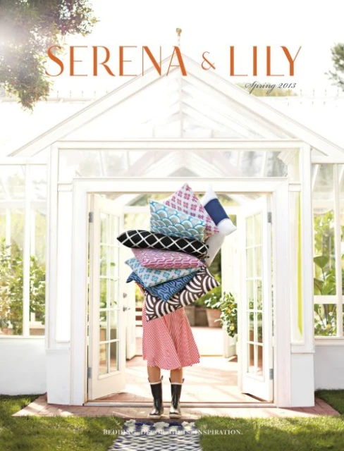 Serena & Lily catalog cover with a woman in a red dress holding a stack of patterned pillows in front of a greenhouse.