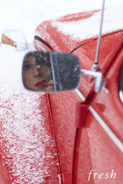 Reflection of a woman applying lipstick in a car side mirror covered in snow next to a red vehicle after winter snowfall.