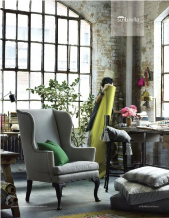 Interior with gray wingback chair, green pillow, patterned cushions, and large window in a sunlit room featuring brick walls.