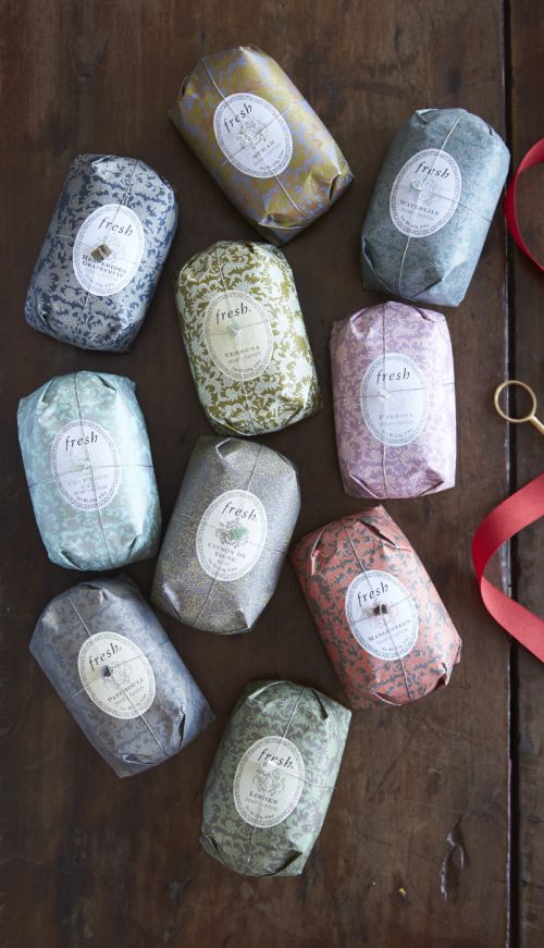 Overhead studio shot of ten colorful, individually wrapped Fresh brand soaps on a wooden surface, accented with scissors and red ribbon.