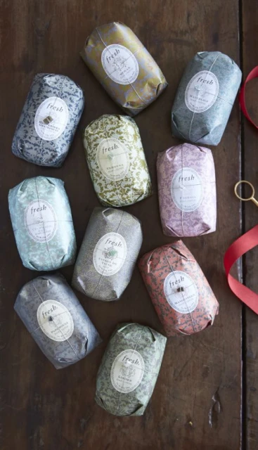 Overhead studio shot of ten colorful, individually wrapped Fresh brand soaps on a wooden surface, accented with scissors and red ribbon.