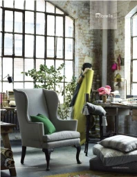 Light grey wingback armchair with green accent pillow in a sunlit industrial loft space with large factory windows.