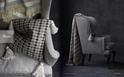 Close-up of a grey armchair with a houndstooth patterned pillow, fringed throw blanket, and tassel details against a dark textured wall