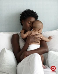 Black woman in white clothes holding an infant child against her chest on a bed