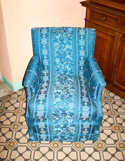 Upholstered turquoise armchair with floral design sits on patterned tile floor, with wood cabinet and pink wall in the background.