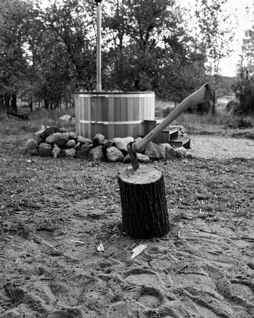 Black and white outdoor photo of a wooden hot tub with an axe stuck in a tree stump in the foreground with a forest background.