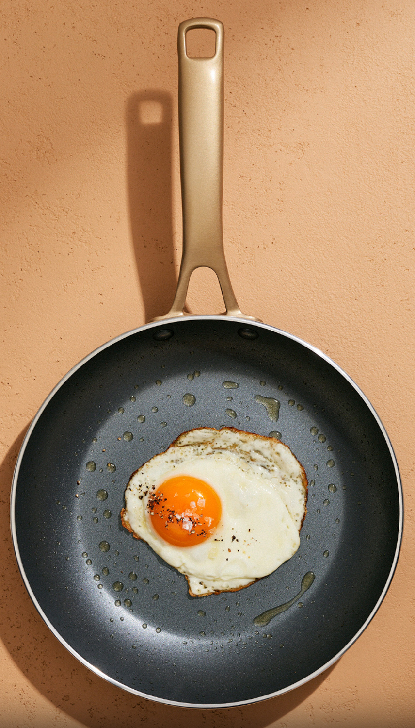 Overhead shot of a fried egg in a non-stick pan with a gold-tone handle, set against a peach-colored textured background. Food photography.