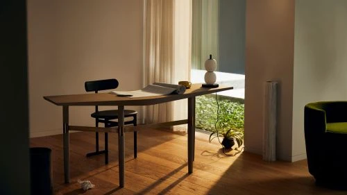 Minimalist home office setup with a wooden desk, black chair, paper, and a modern white lamp, creating a clean and bright workspace.