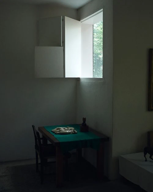 Interior room featuring a wooden table under a window with white shutters, with sculpture and items laid on green cloth.