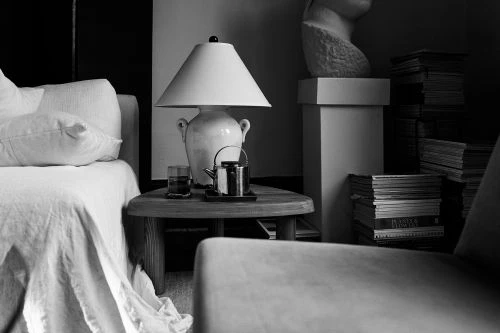 Monochromatic interior with bed, side table, lamp, books, and sculpture of a human head in a dimly lit space.