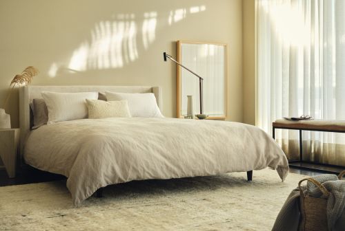 Minimalist modern bedroom interior with a made bed, neutral bedding, sheer curtains, and warm natural sunlight