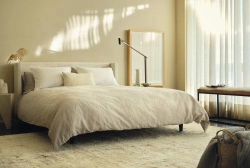 Minimalist modern bedroom interior with a made bed, neutral bedding, sheer curtains, and warm natural sunlight