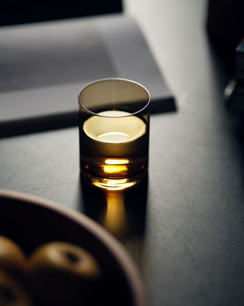 Still life shot of a clear glass on a dark surface, with a bowl and papers blurred in the background, neutral tones, soft lighting.