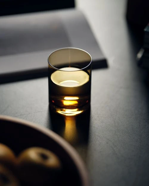 Still life shot of a clear glass on a dark surface, with a bowl and papers blurred in the background, neutral tones, soft lighting.