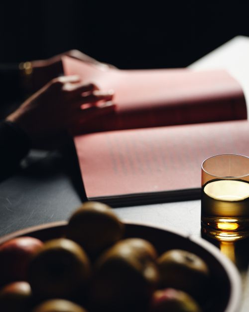 Person reading a book at a table indoors next to a glass of yellow liquid and bowl of fruit; soft lighting creates a relaxing ambiance.