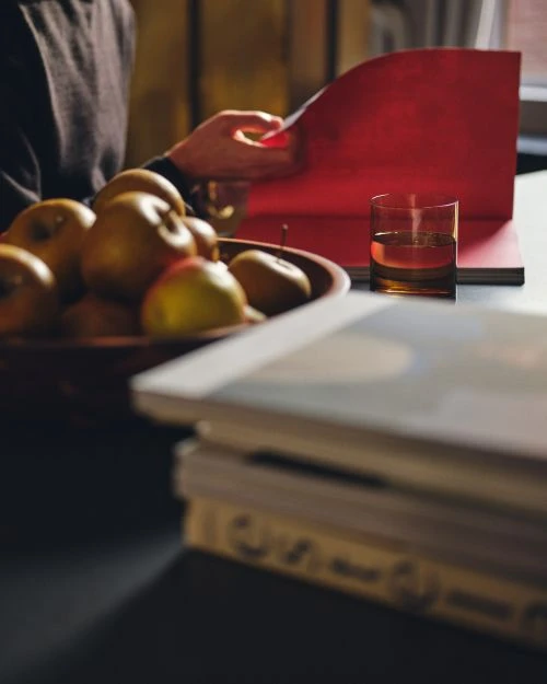 Still life photo of a wooden bowl filled with apples, open red notebook, and glass of whiskey, set on a table in a warm indoor setting.