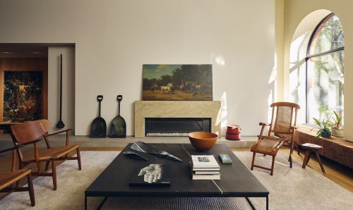 Modern living room interior featuring a black coffee table, wooden chairs, a fireplace with artwork, and a large arched window.