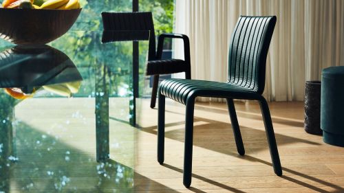 Interior shot of two dark green chairs on a wood floor, with glass table and fruit bowl in the background, bathed in natural light.