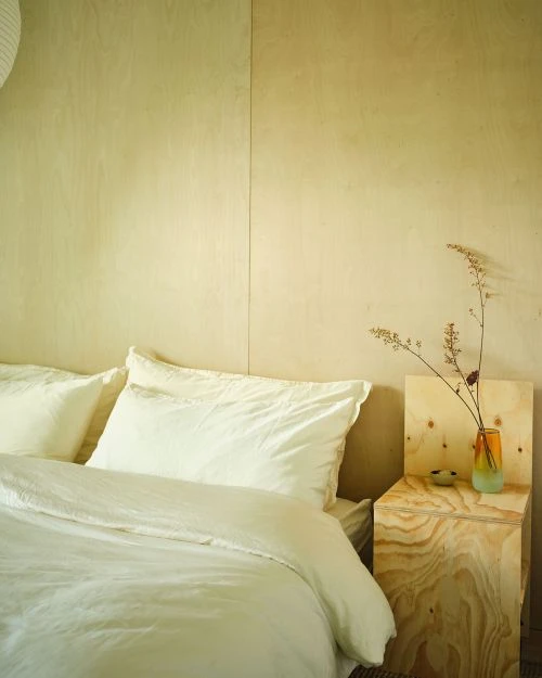 Minimalist bedroom interior featuring a bed with white linens, a wooden bedside table with a vase, and light wood paneling.