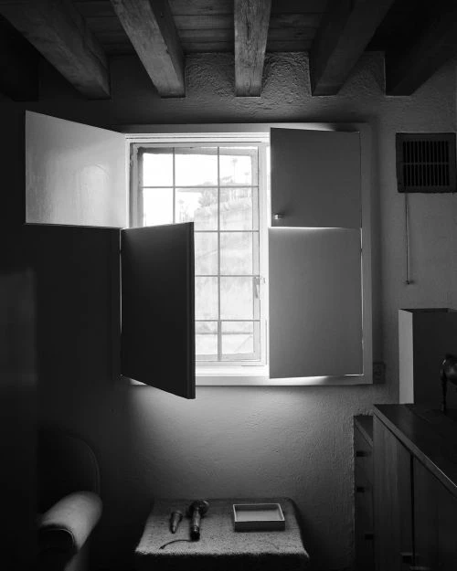 Black and white interior shot of a window with open shutters, a table with objects, and rustic wood ceiling beams, emphasizing light and shadow.