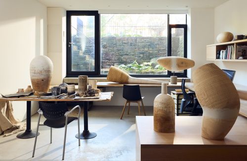 Interior photo of an art studio featuring sculptural wooden art pieces, tables, chairs, natural light, and a view of the garden.