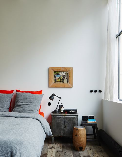 Modern bedroom interior featuring a gray bed with orange accents, a rustic metal nightstand, and a framed picture on a white wall.
