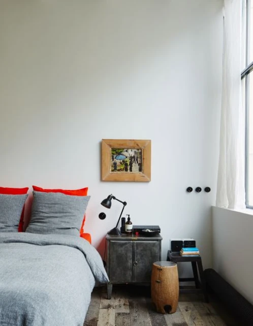 Modern bedroom interior featuring a gray bed with orange accents, a rustic metal nightstand, and a framed picture on a white wall.