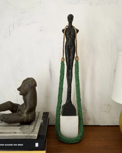 Bronze statue of a nude male figure stands on a white base with a green beaded necklace alongside a book stack with a bronze figure.