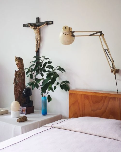 Bedroom still life of crucifix, religious figures, plant vase, lamp, and bed create a peaceful, spiritual, and restful interior scene.