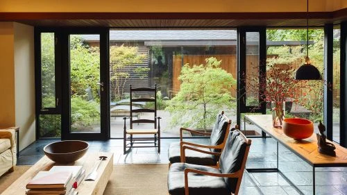 Living room featuring modern interior with minimalist decor, stylish armchairs, wooden table and bowl, and large windows overlooking the garden.