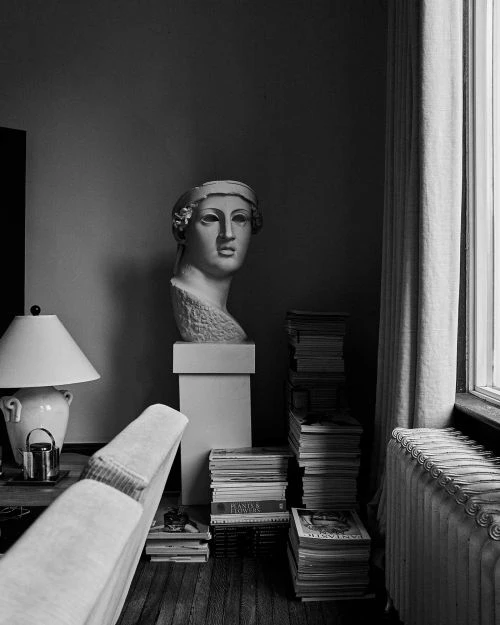 Monochrome indoor scene with a neoclassical-style bust, stacks of books, a table lamp, and radiator near a window with curtains.